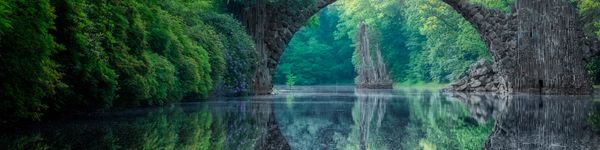 A serene stone bridge arches over a reflective river surrounded by lush greenery.