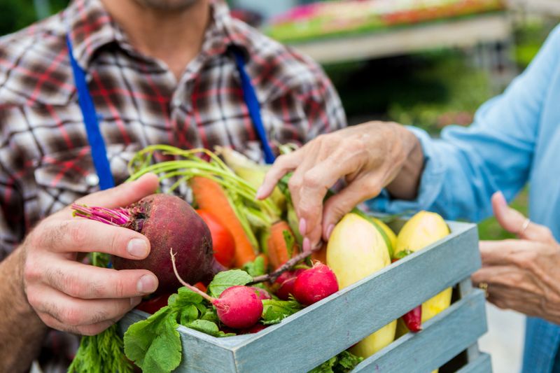 Male farmer selling fresh summer vegetables to senior woman. Carrots, radishes,  and a beet are all in the small basket. The man is wearing a blue apron. Close up of vegetables and hands, the faces are out of the shot.