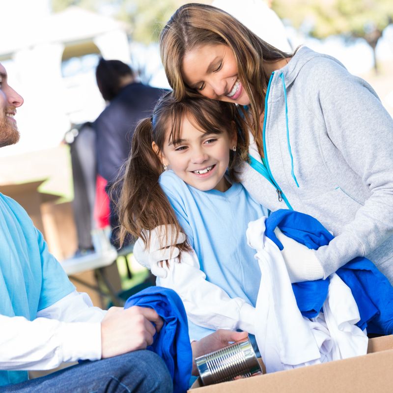 Happy mother hugs her elementary age daughter as they receive clothing at outdoor community clothing drive. The father is sitting beside them smiling. They are pulling items of clothing and canned goods out of a box. Volunteers are working in the background. It is a bright sunny day.