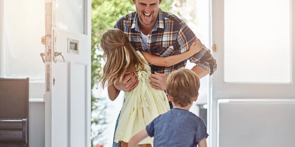 Father joyfully embraces his daughter at the front door as son runs towards them.