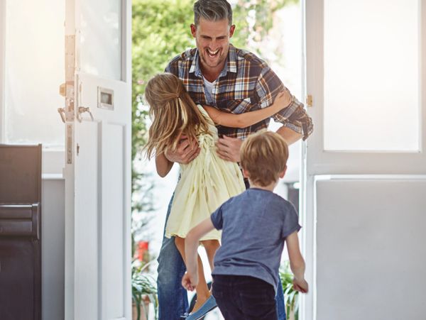 Father joyfully embraces daughter while son runs toward them at the door.