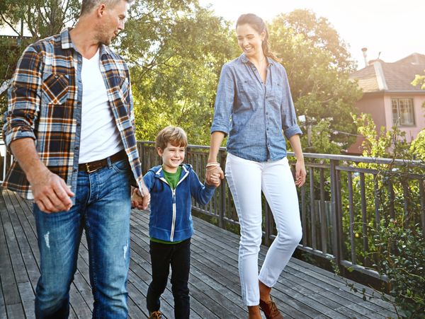 A happy family of three walking hand in hand outdoors on a wooden deck.