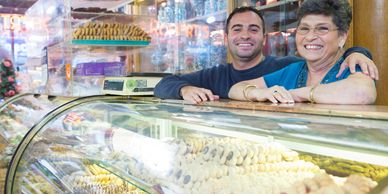 Smiling bakery owners posing behind a glass display filled with cookies and pastries.