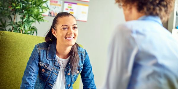 A woman in a denim jacket smiling during a conversation with another person.