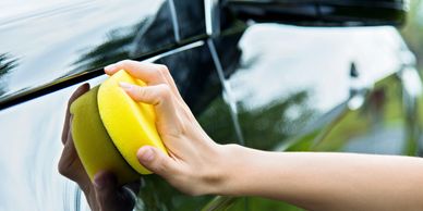 Hand washing a black car with a yellow sponge.