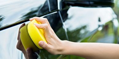 Hand polishing a black car with a yellow sponge.
