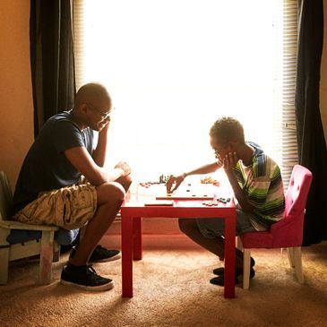 Two people intensely playing chess at a small red table by a bright window.