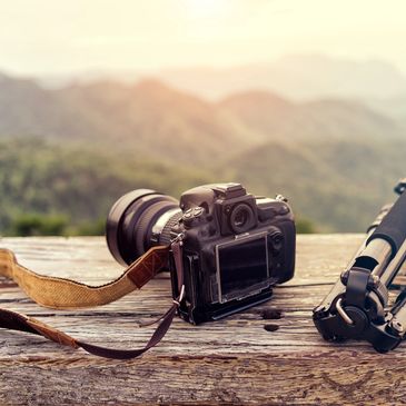 A DSLR camera and tripod resting on a wooden surface with mountains in the background.