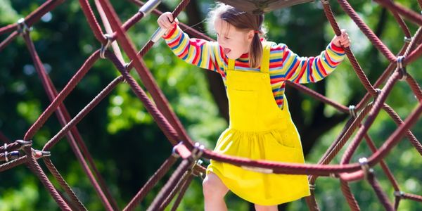 A girl in a yellow dress plays on a rope climbing structure outdoors.