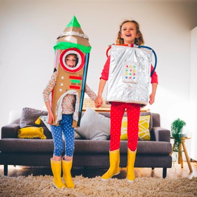 Two children dressed in homemade rocket and robot costumes, holding hands indoors.