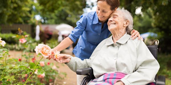 A caregiver and elderly woman share a joyful moment outdoors among flowers.