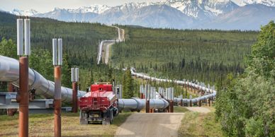 A long pipeline winds through a forested area with mountains in the background.