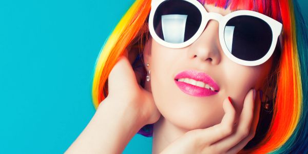 Woman with vibrant rainbow hair and white sunglasses against blue background.