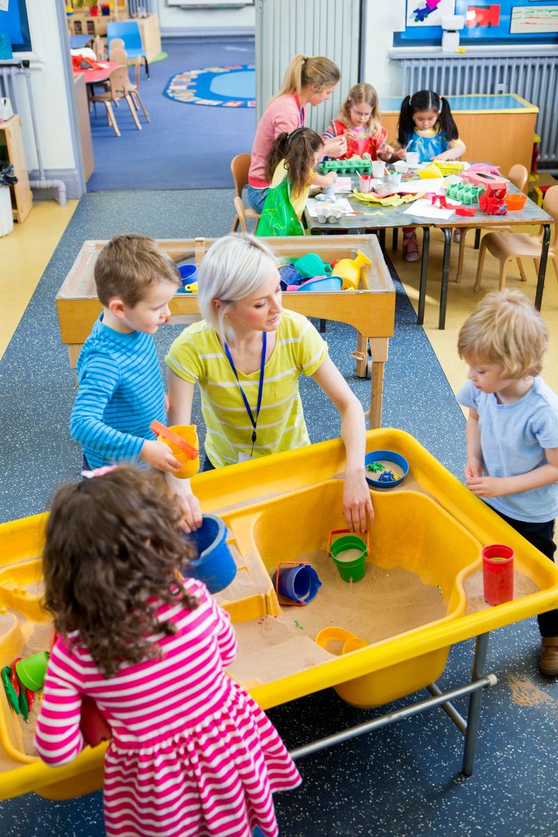 Elevated view of teachers doing activities with children in nursery. Some are playing in a sand box and the others are making crafts.
