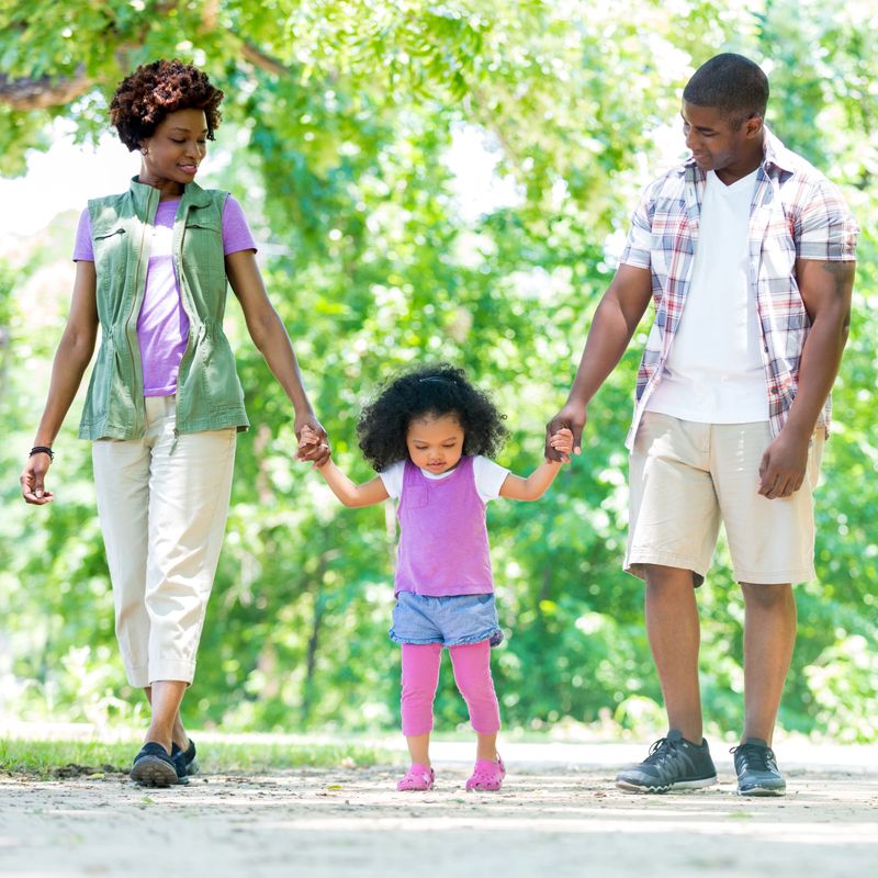 Loving African American mid adult parents tenderly hold their toddler daughter's hands as they walk down path in the park. They look at the little girl as they walk. They are dressed in trendy casual attire. Trees surround them.