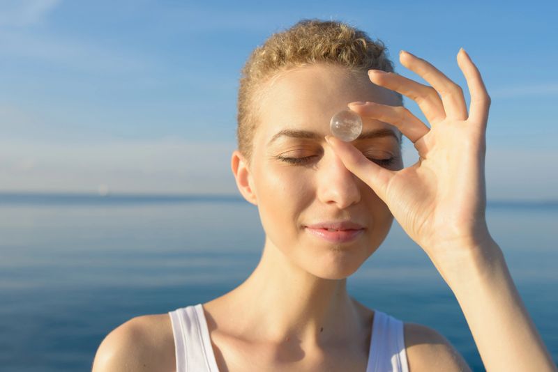 A young woman holds a quartz crystal sphere on her forehead; standing by the sea at dawn, Antibes Juan-les-Pins, Cote d'Azur, France.