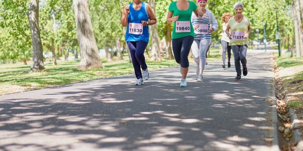 Women of different ages joyfully running a marathon in a tree-lined park.