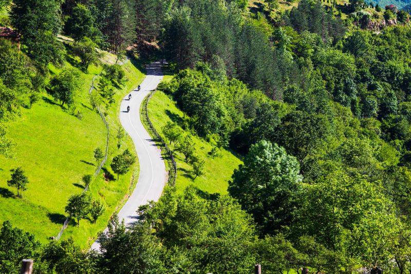 Aerial view of three motorcyclists on a winding rural road through a scenic lush green forested mountain valley
