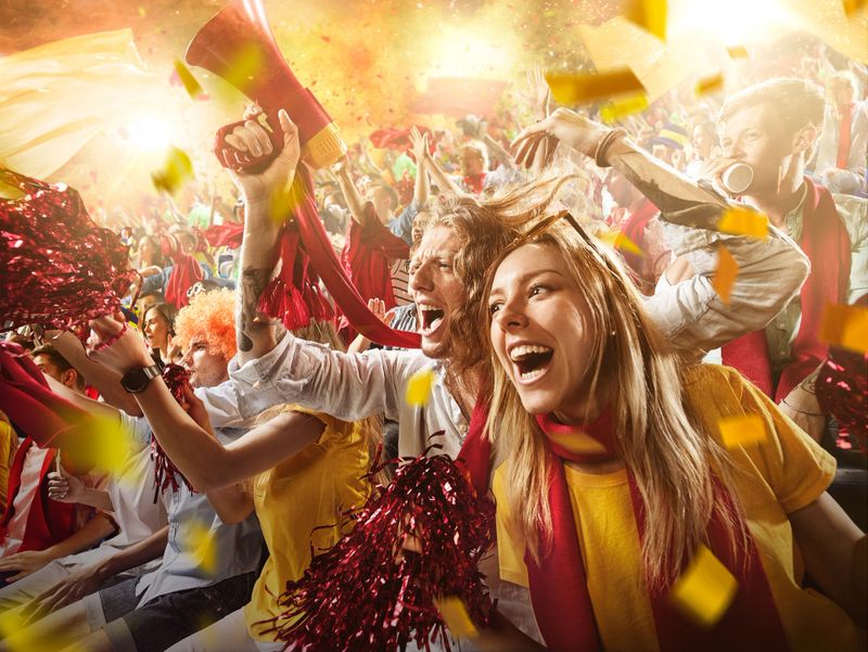 :biggrin:On the foreground a group of cheering fans watch a sport championship on stadium. Everybody are happy. People are dressed in casual cloth. Colourful confetti flies int the air.