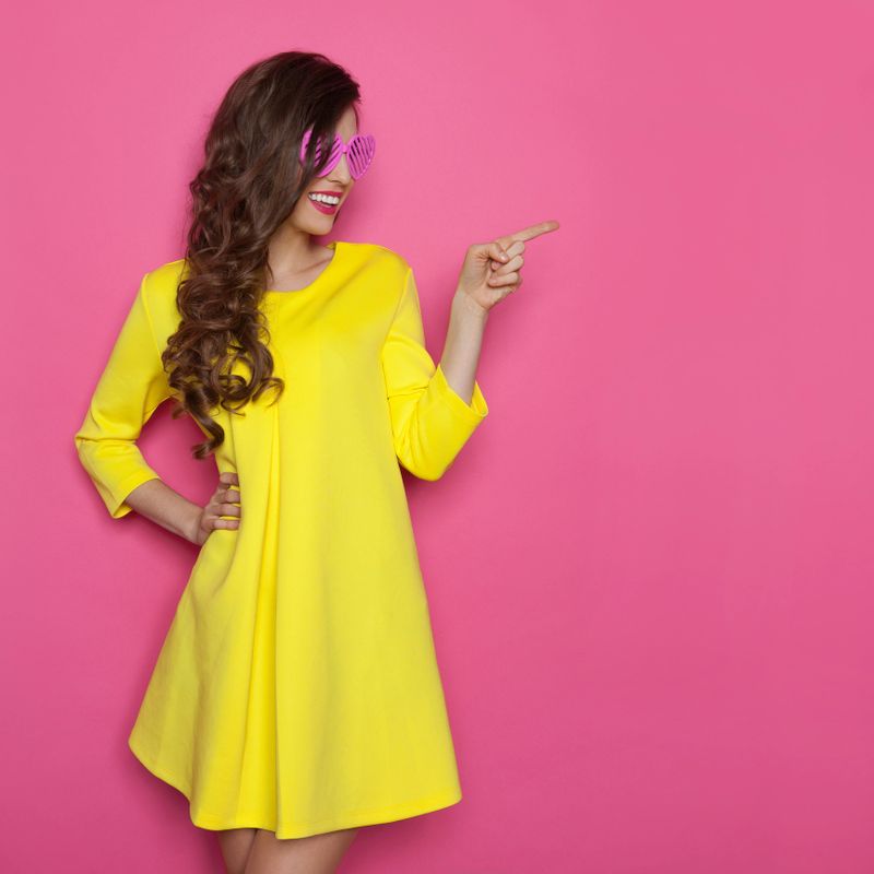Smiling beautiful young woman with curly long brown hair  posing in yellow dress with hand on hip, pointing and looking away. Three quarter length studio shot on pink background.