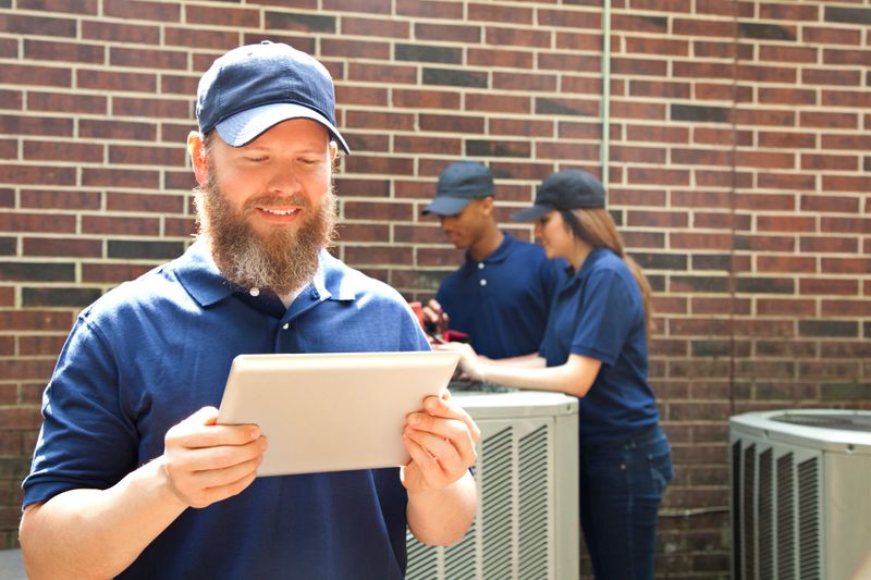 Multi-ethnic group of two men and one woman repairing a home's air conditioner unit outdoors. Mid-adult man in foreground uses a digital tablet to access the repairs.  They all wear blue uniforms.