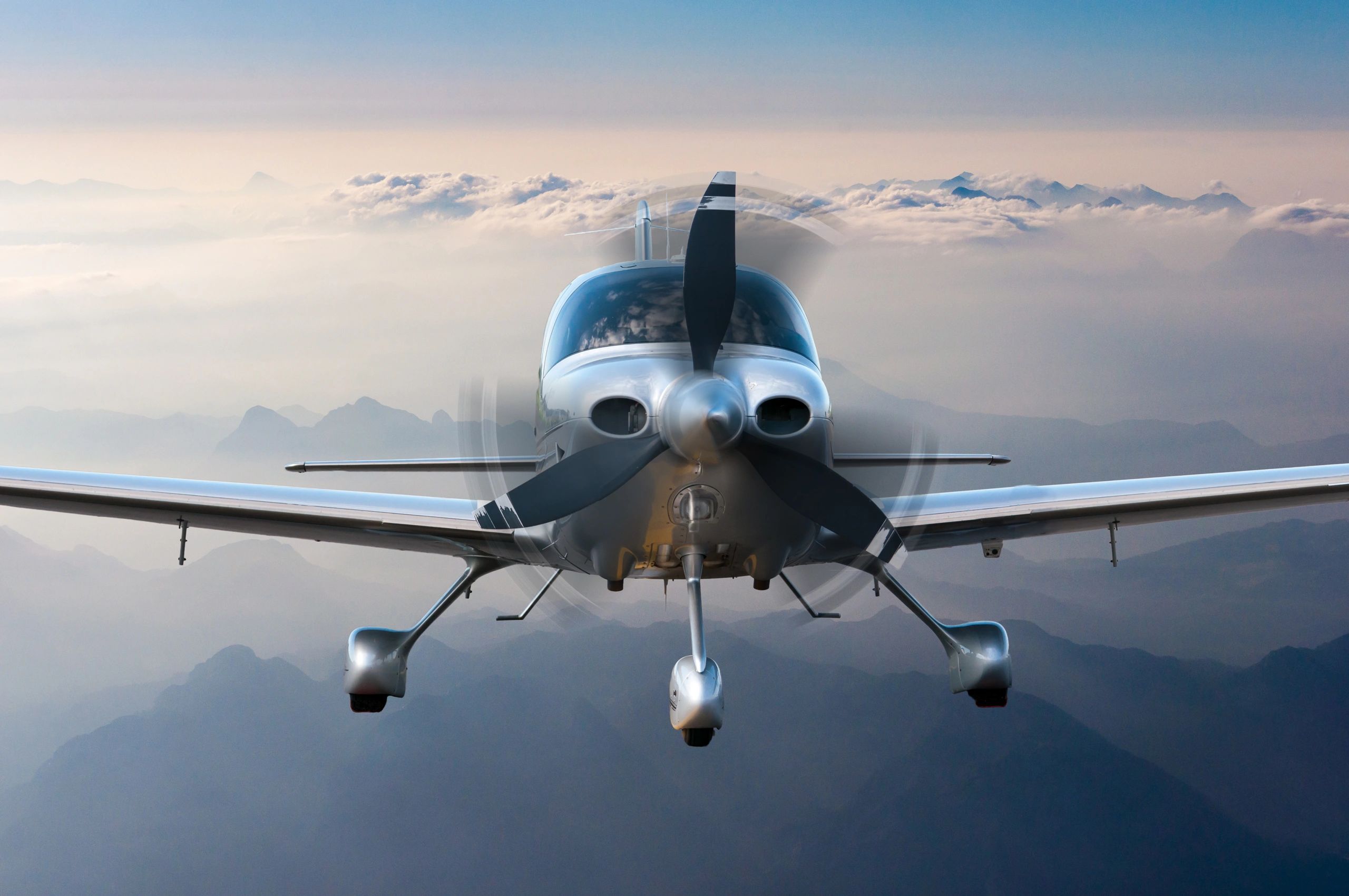 Front view of a small propeller airplane flying above mountains with clouds.
