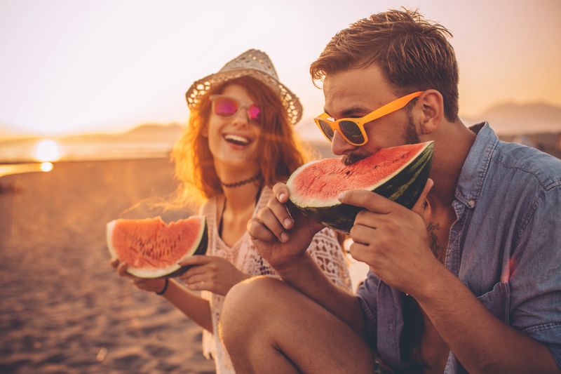 Hipster couple sitting on the beach and eating watermelon. Wearing casual summer clothing, hat and sunglasses. Enjoying in sunset by the sea.