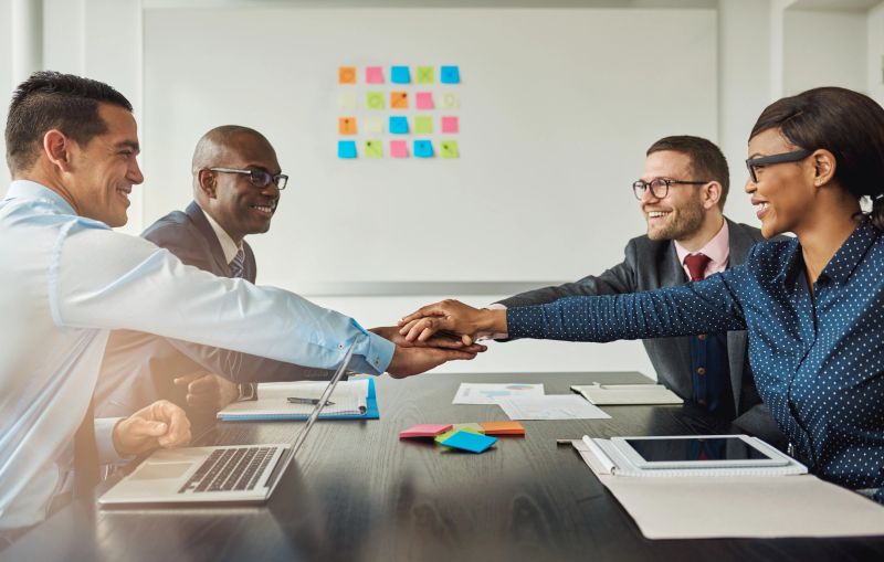 Multiracial business team signalling their commitment to each other by reaching across the table in the office to stack hands as they smile at each other