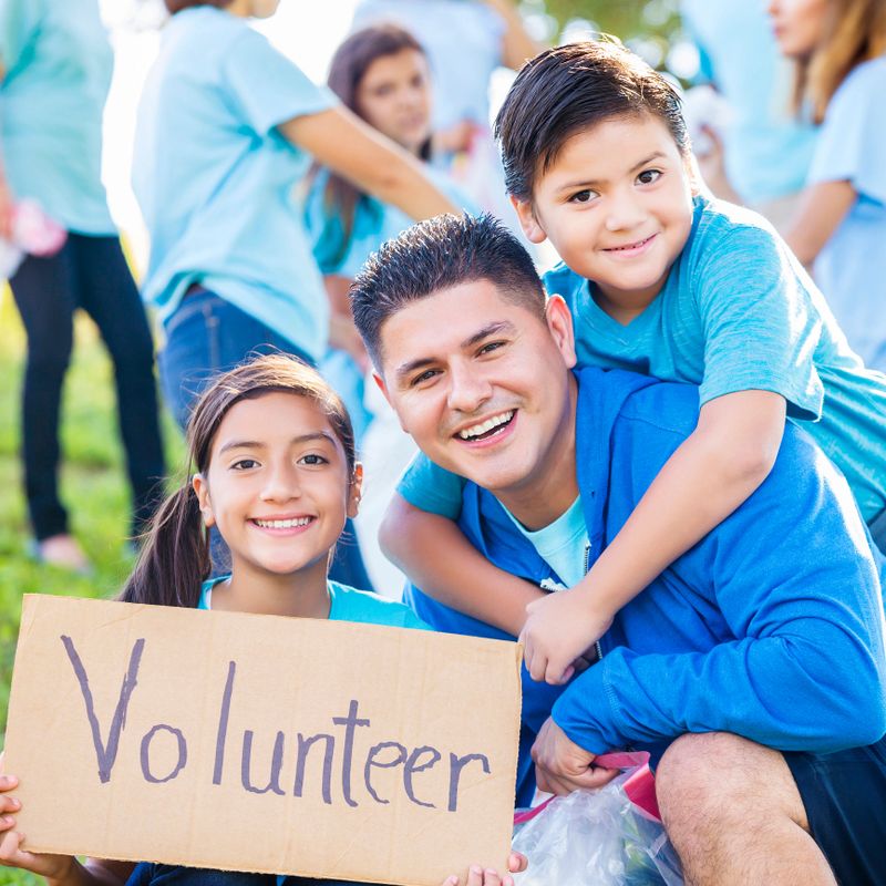 Mid adult Hispanic dad and his elementary age son and preteen daughter help neighbors clean up their park. The girl is holding a cardboard sign with "Volunteer' printed on it. They are wearing blue t-shirts and the dad has a royal blue hoodie over his t-shirt. Their friends are talking in the background.