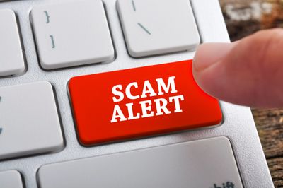 Finger about to press a red keyboard key labeled 'SCAM ALERT'.