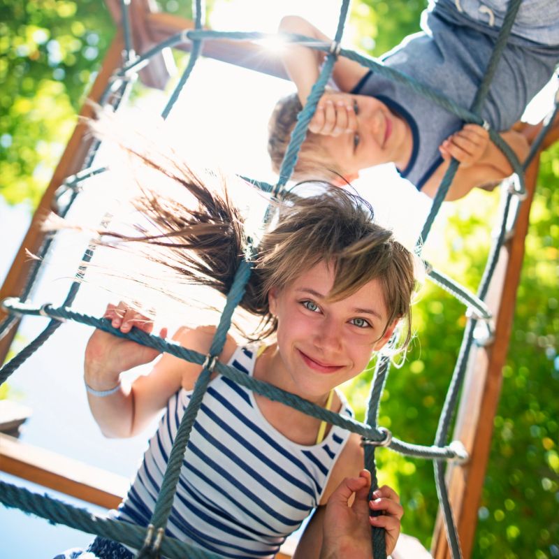 Portrait of little girl climbing in the playground. The girl aged 10 is smiling at the camera and her brother aged 6 is smiling in the background. Sunny summer day. Shot from directly below the kids.
