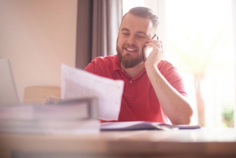 A young male possibly a single parent or small business owner sits at home talking to the bank or finance company on the phone. He is paying off his remaining debt and is feeling very happy about it. He is smiling as he listens to the advisor.