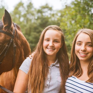 Two smiling girls with a brown horse outdoors.