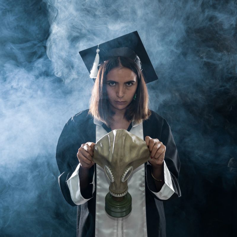 Newly graduate female journalist holding gas mask on her face.She is in fog and looking at camera. Shot in studio with medium format camera Hasselblad.Cropped to square framing.Gas mask and fog are used since journalists in many countries around the world have to wear gas mask in order to photograph protests.