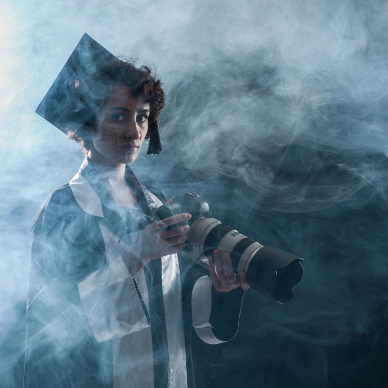 Newly graduate female photo journalist holding camera while she has yellow crime scene tape on her gown.She is in fog and looking at the camera. The background is black.Shot in studio with medium format camera Hasselblad.Cropped to square framing.Gas mask and fog are used since journalists in many countries around the world have to wear gas mask in order to photograph protests.