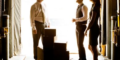 Three business professionals discuss logistics near stacked boxes at a warehouse loading dock.