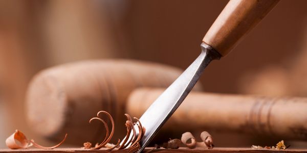 Wood carving chisel shaping fine curls of wood with a mallet in the background.