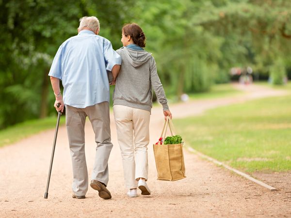 Elderly man and woman walking arm in arm on a path, carrying groceries.