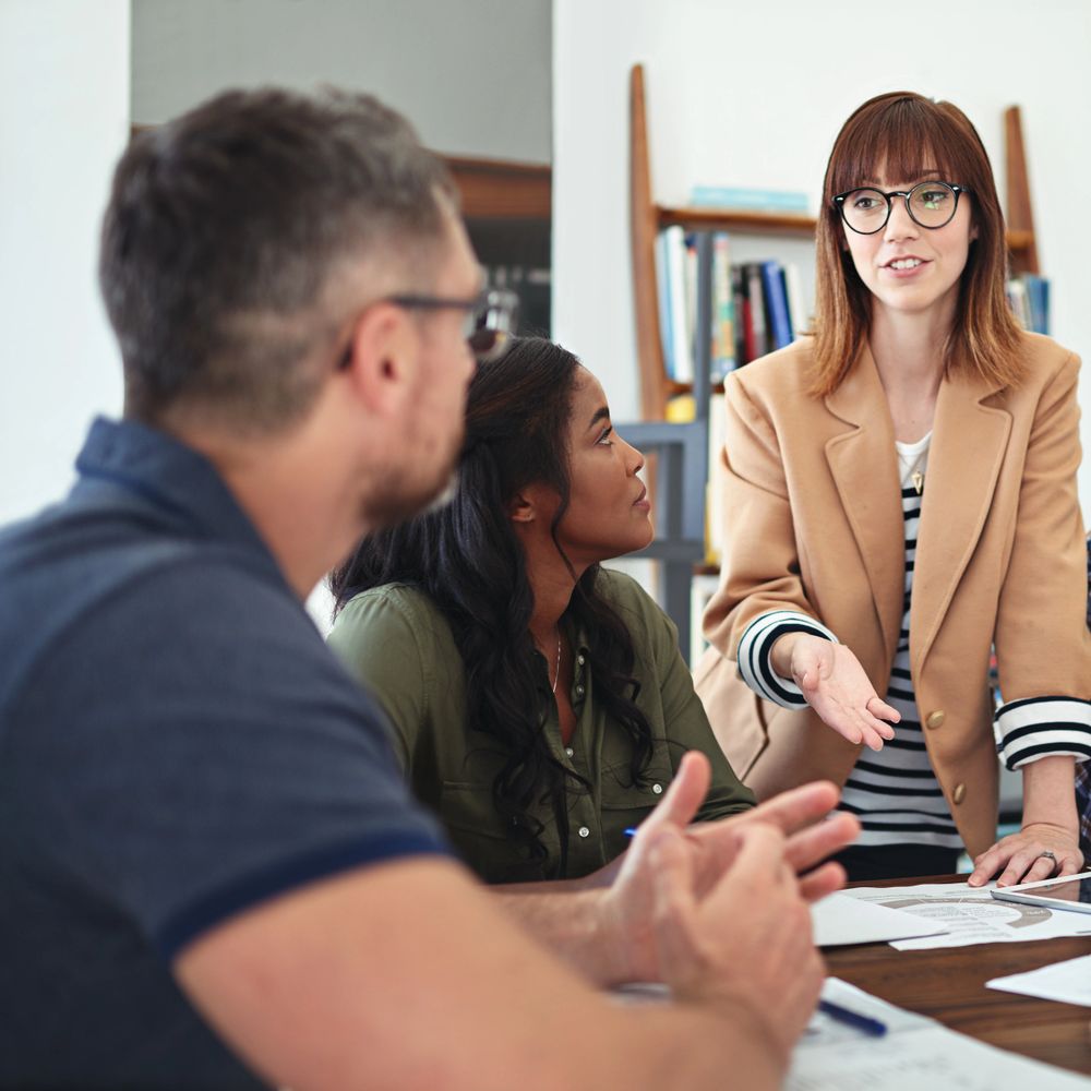 A woman in glasses leads a discussion with two colleagues in an office setting.