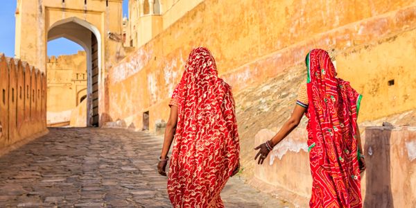 Two women in vibrant red traditional attire walking up a stone path near a historic fort.