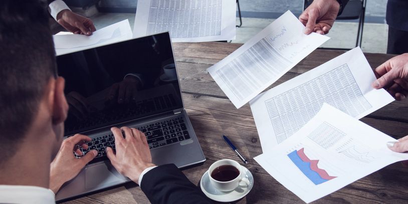Business professionals discussing data charts and reports around a laptop.