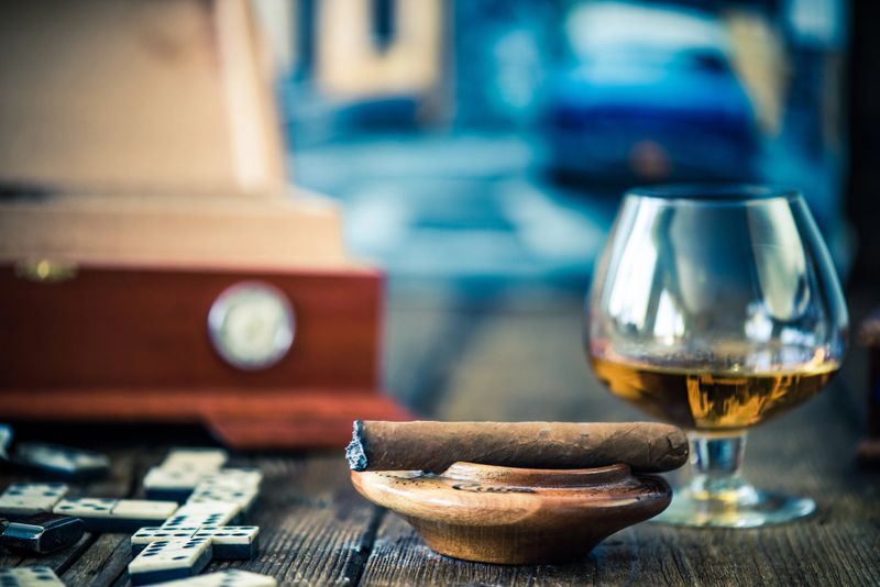 cuban cigar and glass of rum or cognac on wooden table with cuban painting in background