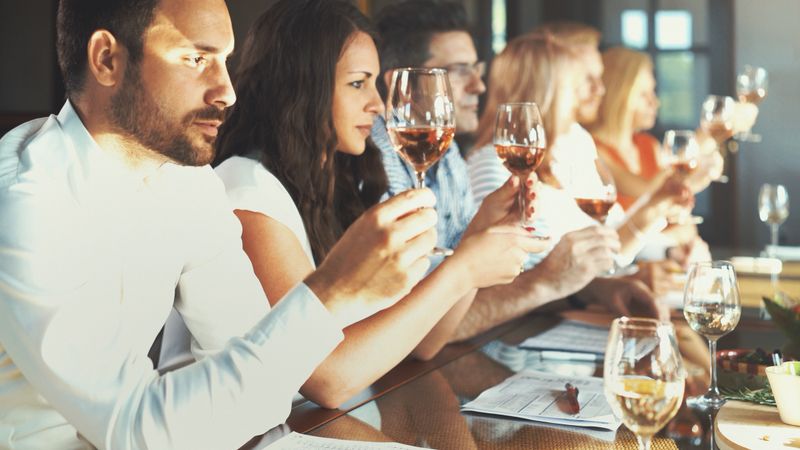Closeup side view of group mixed age adults tasting different types of wine at a winery. They are seated by a long table with wines in front of each person and wine evaluation charts. Focus on the closest person to the camera.