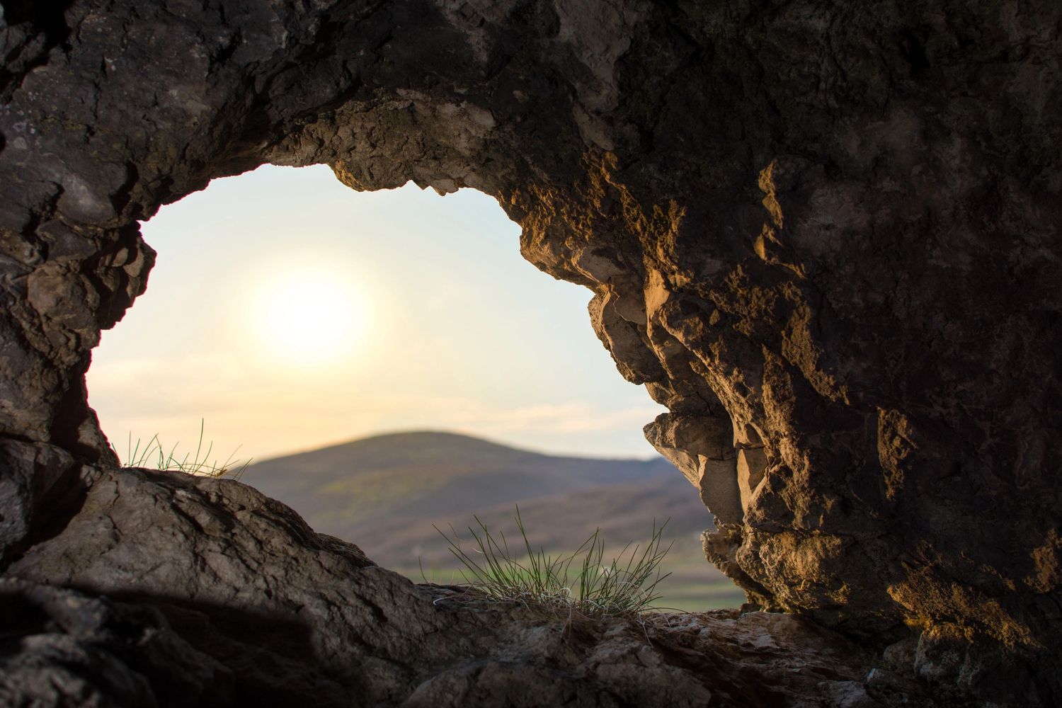 Sunset view through a rocky cave opening with distant hills.