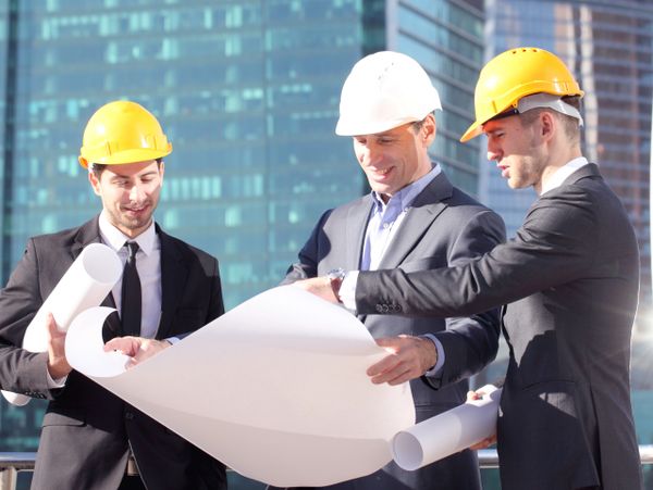 Three architects in suits and helmets reviewing blueprints outdoors.