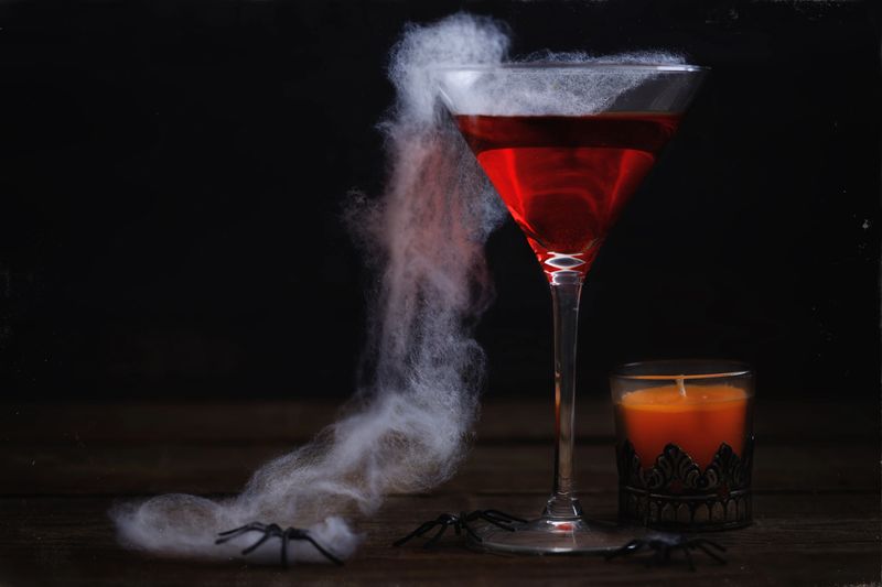 Spooky Halloween drink, spider web and orange candle over rustic wooden background. Aging effect, colour toning, atmospheric noise and scratches