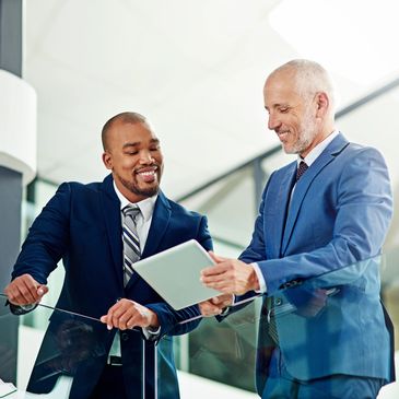 Two businessmen in suits smiling and discussing on a digital tablet.