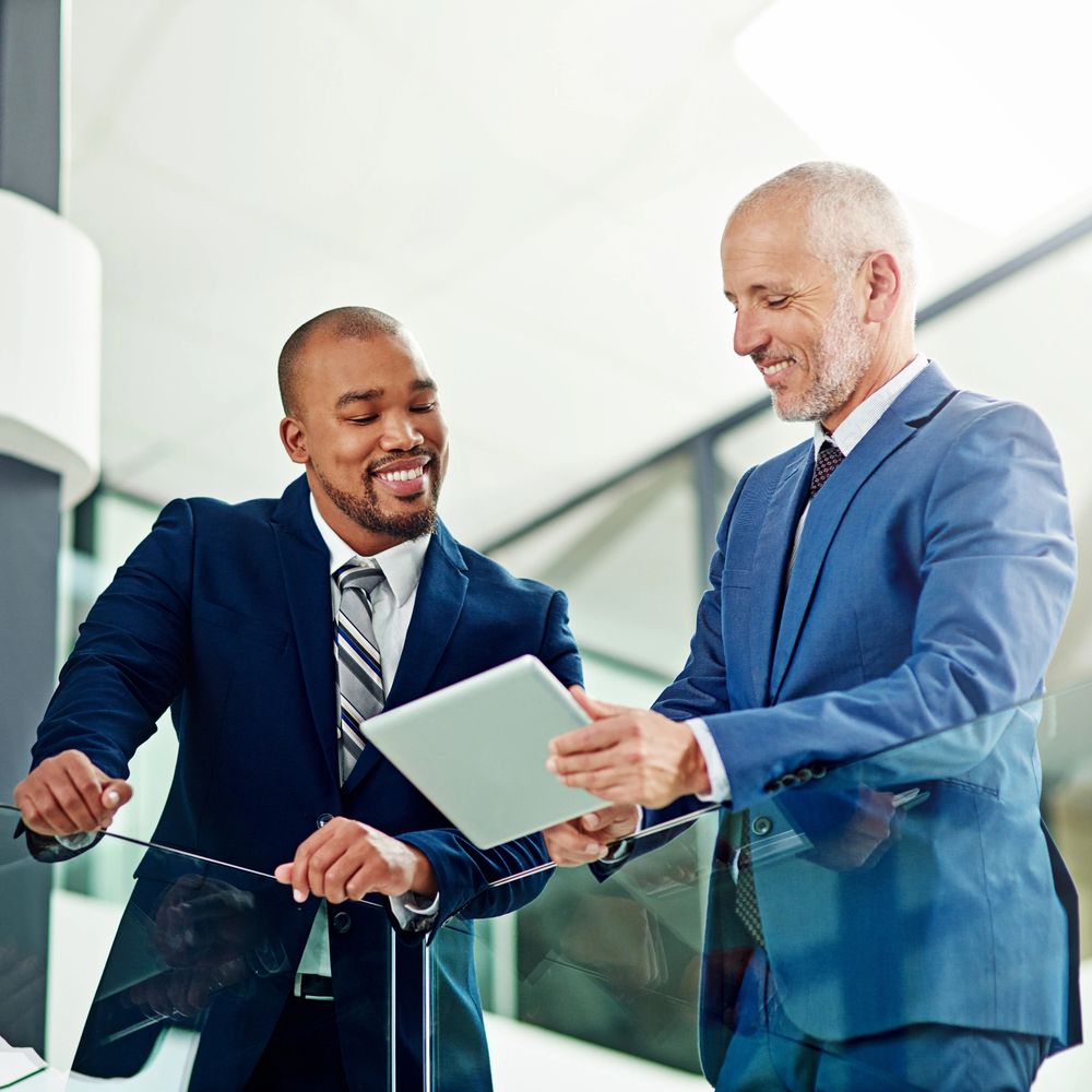 Two businessmen in suits smiling and discussing on a digital tablet.