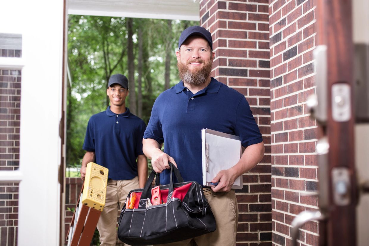 Two repairmen at a doorstep with tools and clipboard.