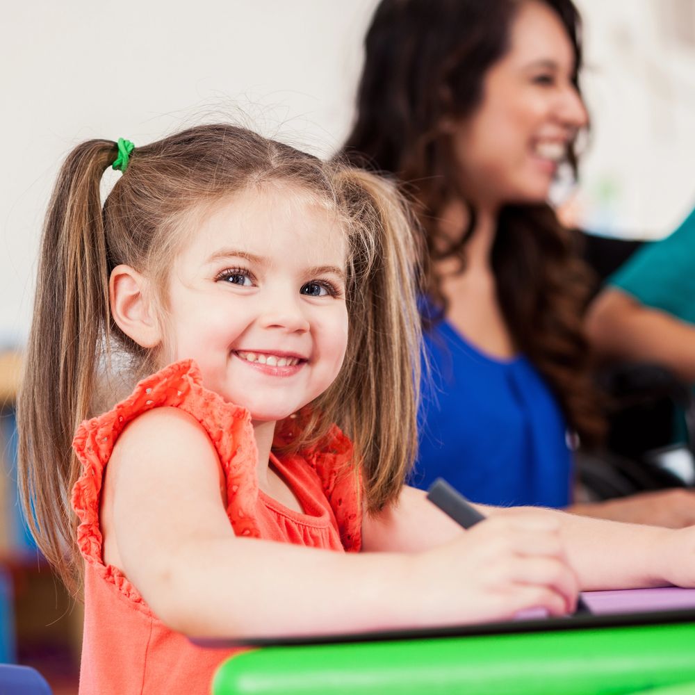 Smiling young girl with pigtails drawing at a table with a woman in the background.